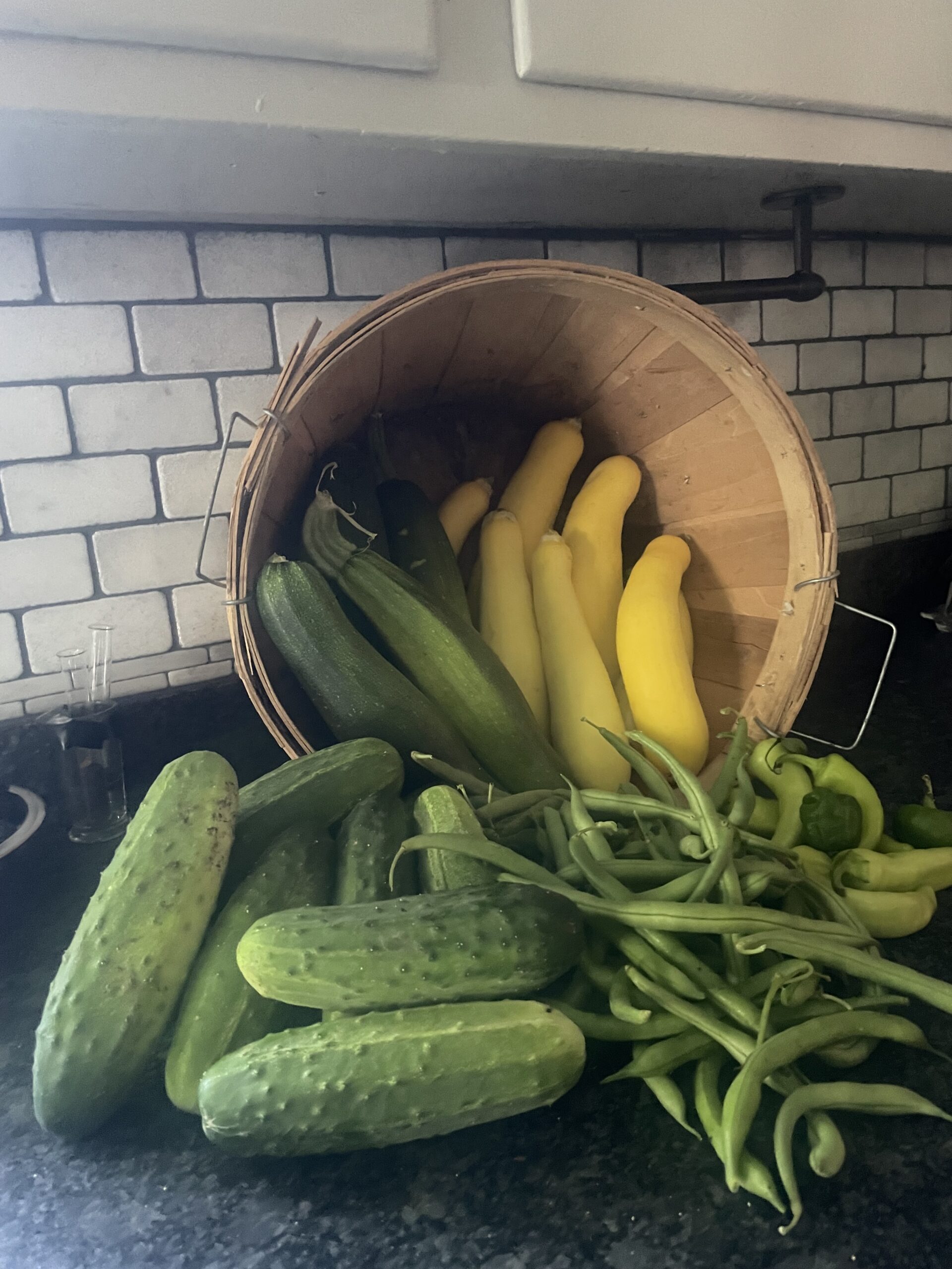 Freshly harvested vegetables including cucumbers, yellow squash, and green beans displayed in a wooden basket on a kitchen countertop.